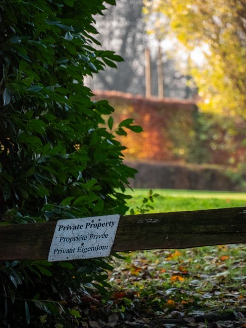 A wooden fence marks the boundary of a private garden, with a white sign attached that reads 'Private Property' in English, French, and German. Behind the fence, there is a lush green lawn bordered by
