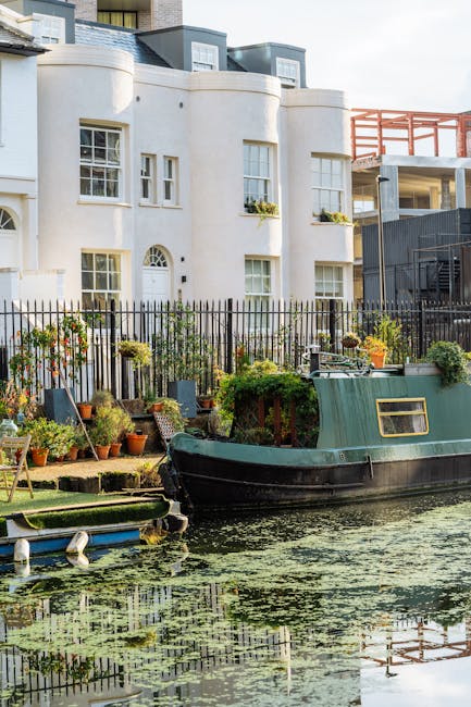 A tranquil canal scene featuring a narrowboat moored alongside a garden filled with potted plants and flowering shrubs in front of a white, curved-fronted building with multiple windows. The garden ar